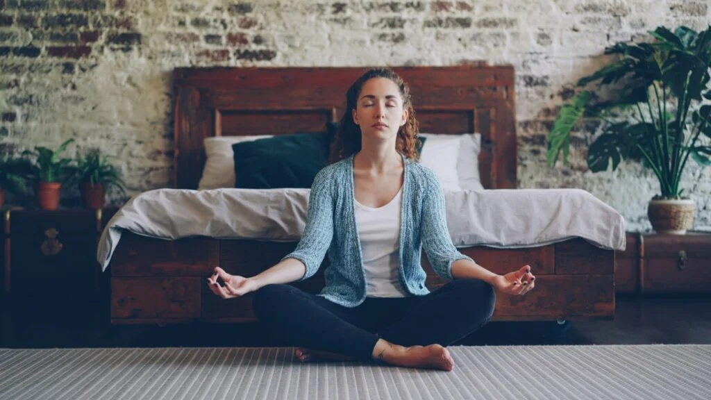 Woman sitting cross-legged in her bedroom with eyes closed, breathing slowly