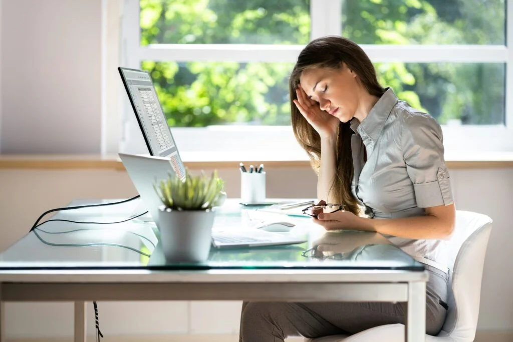A tired woman at her desk in the afternoon, holding her head with her eyes closed
