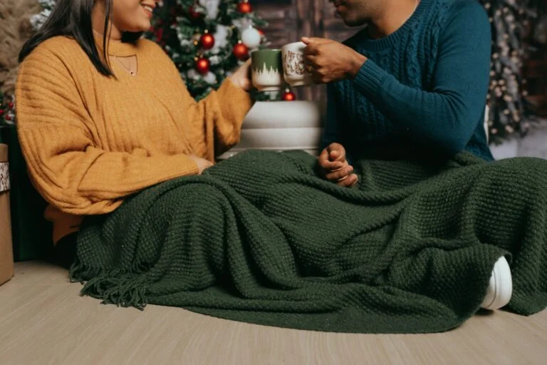 Two people sitting under a green knitted blanket with mugs in front of a Christmas tree