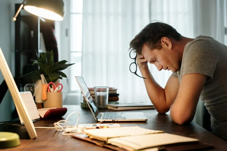 A stressed man at a desk with his head in his hand, holding his glasses, in front of a laptop