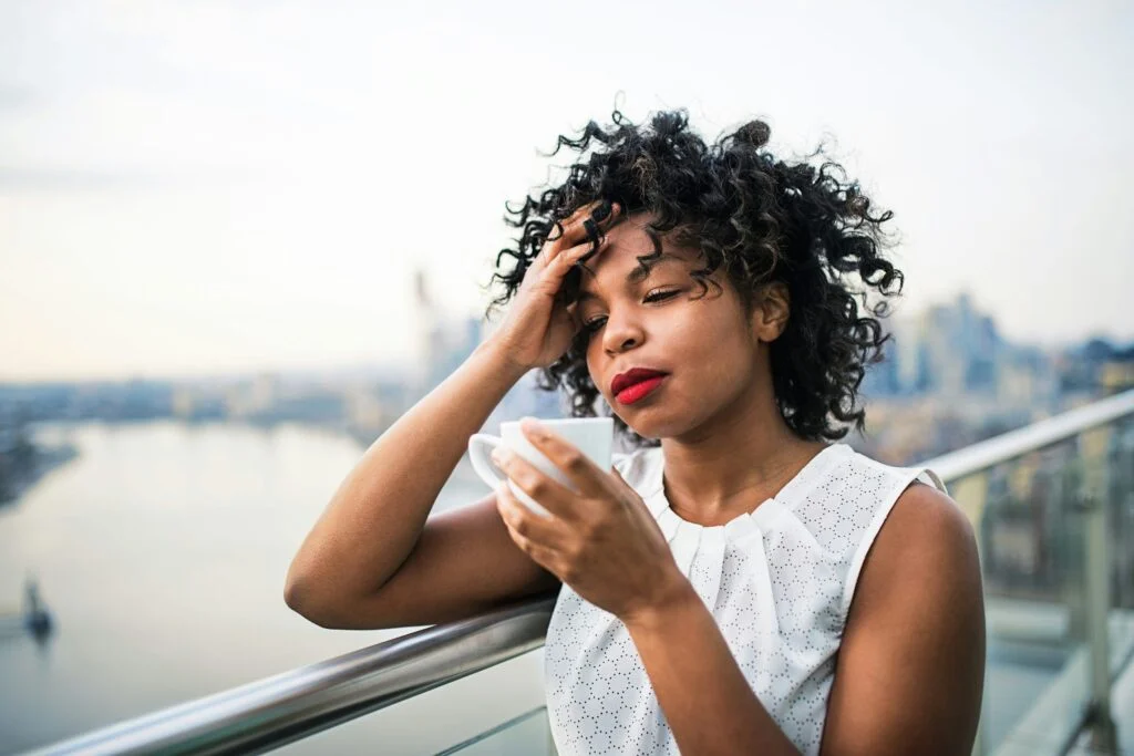 A tired woman holding a cup of coffee on a balcony, with her hand on her head