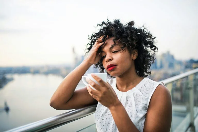 A tired woman holding a cup of coffee on a balcony, with her hand on her head