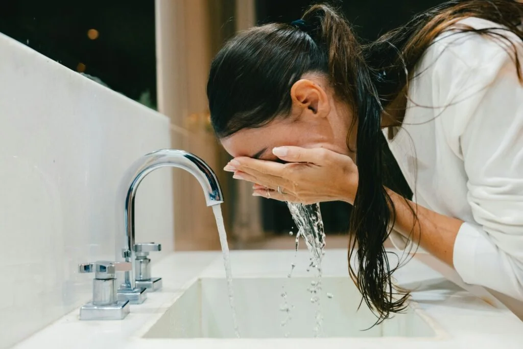 A woman splashing cold water on her face at a bathroom sink