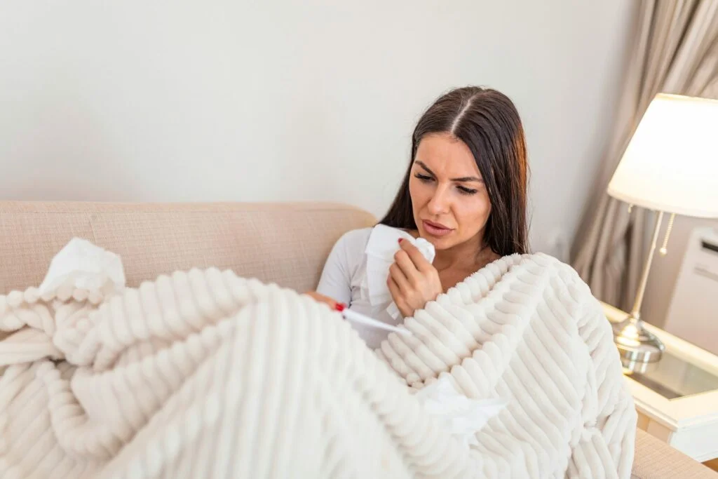 A woman wrapped in a cream blanket on a sofa holding a tissue, with a cold