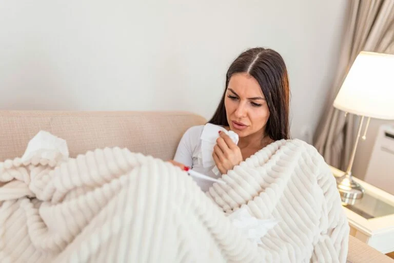 A woman wrapped in a cream blanket on a sofa holding a tissue, with a cold