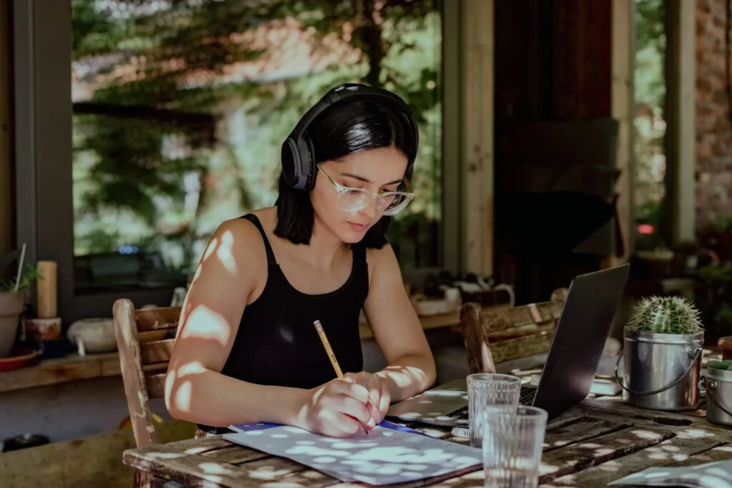 A woman wearing headphones concentrating on writing notes at a sunny table