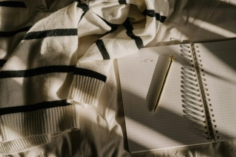 An open notebook with a gold pen next to a striped jumper on a bed in soft sunlight