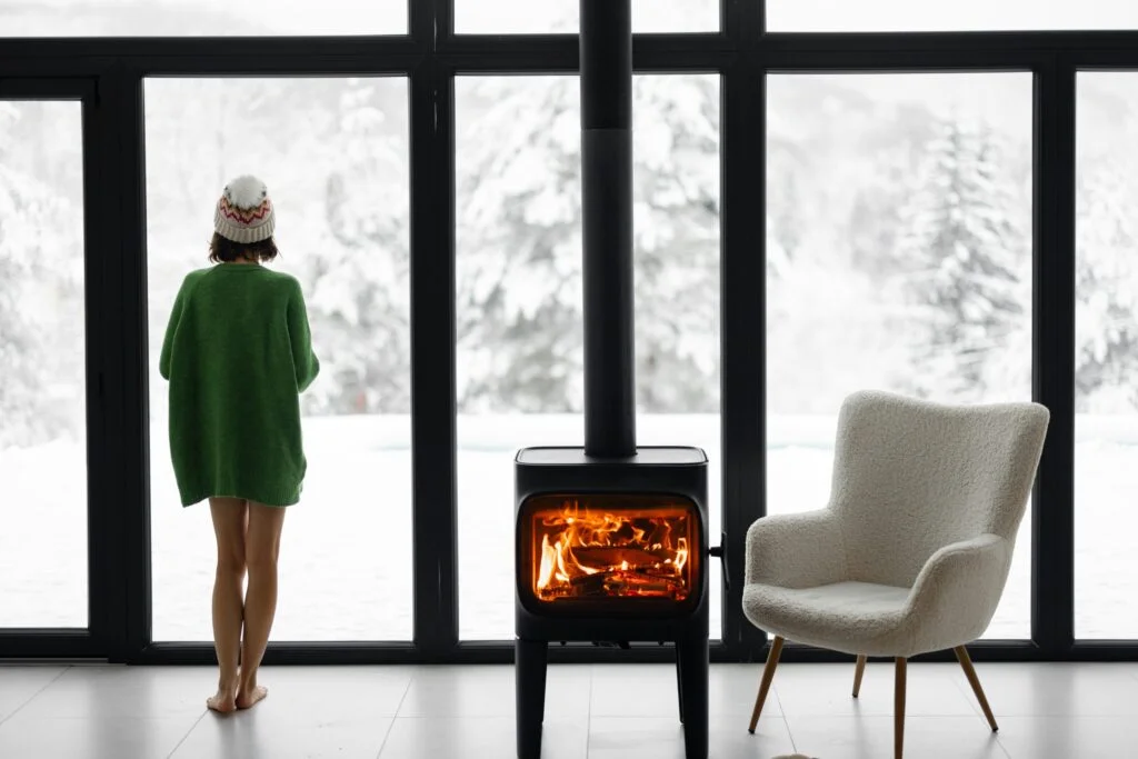 A woman in a green cardigan standing alone at a large window looking out at snow beside a wood burner