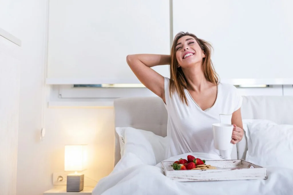 A smiling woman stretching in bed with a cup of coffee and a breakfast tray of strawberries