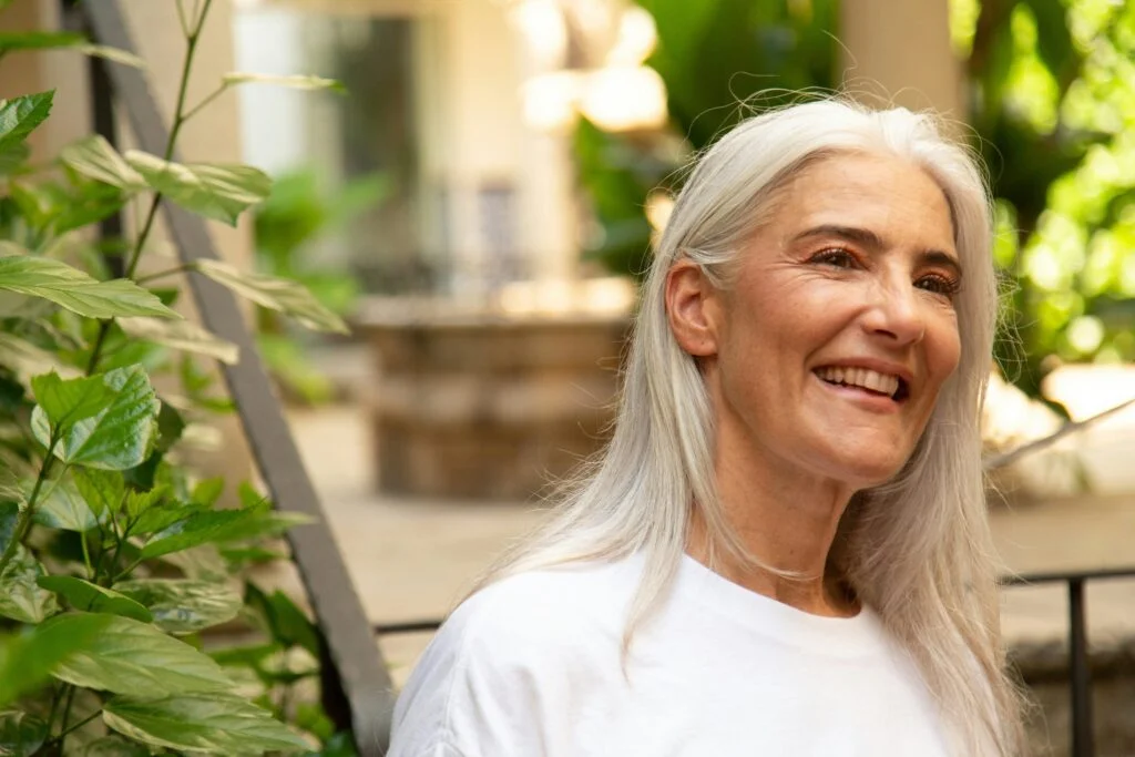 A smiling woman with long grey hair outdoors among green plants