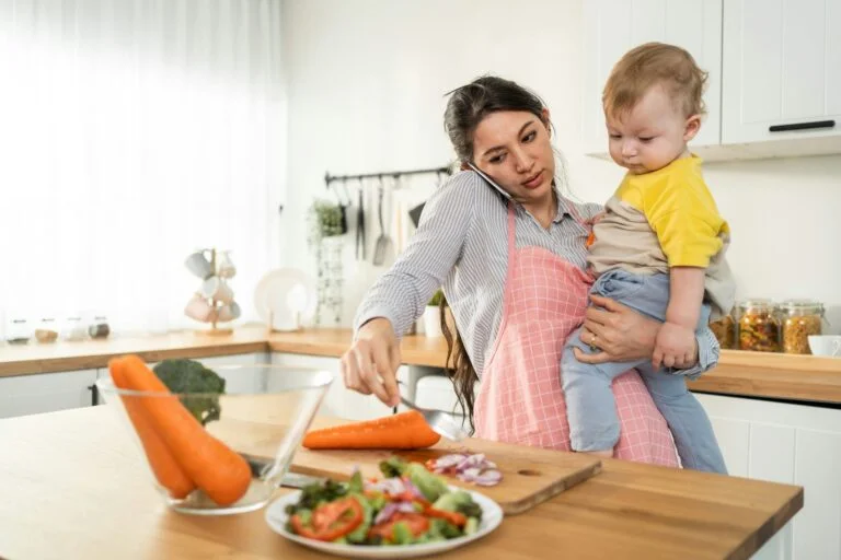 A busy mother on the phone holding her baby while preparing a salad in the kitchen