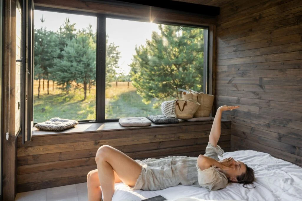 A woman stretching in bed on a sunny morning by a large window looking out on trees