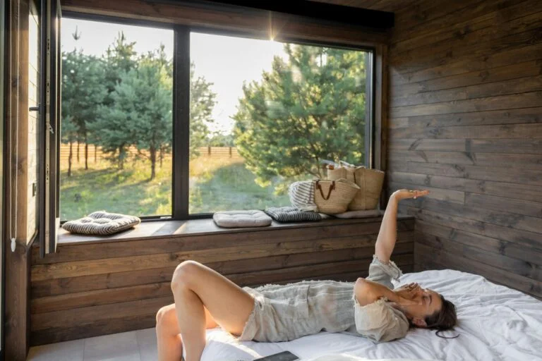 A woman stretching in bed on a sunny morning by a large window looking out on trees