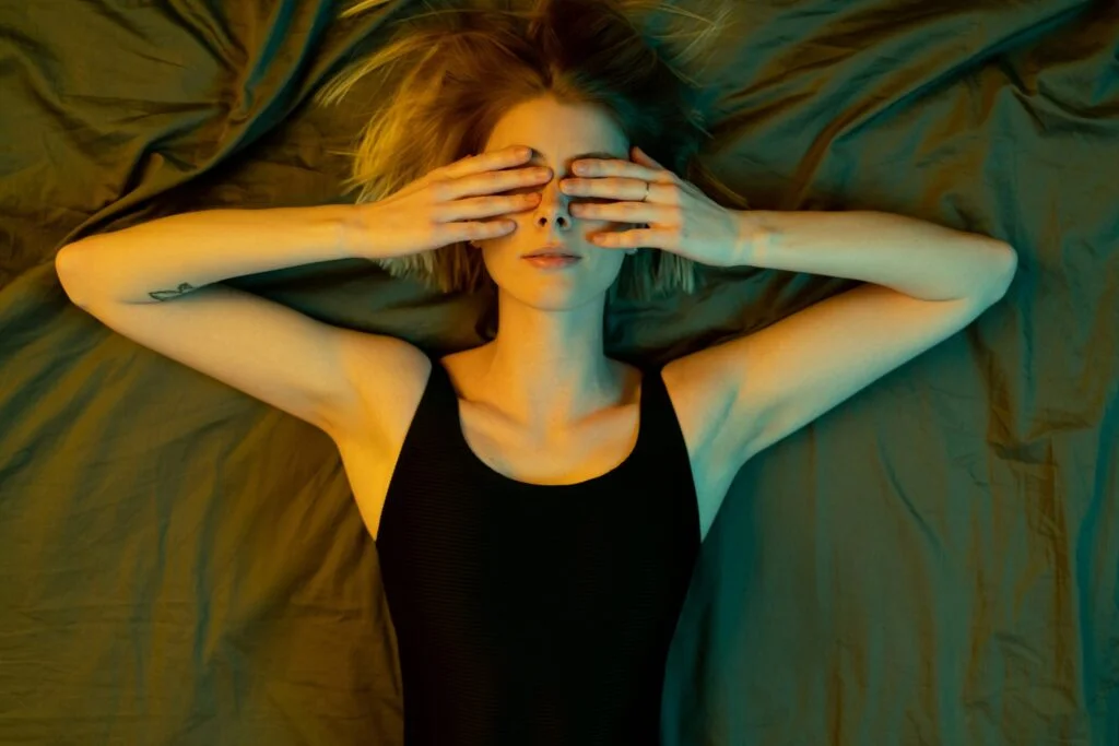 A woman lying in bed covering her eyes with both hands in dim light