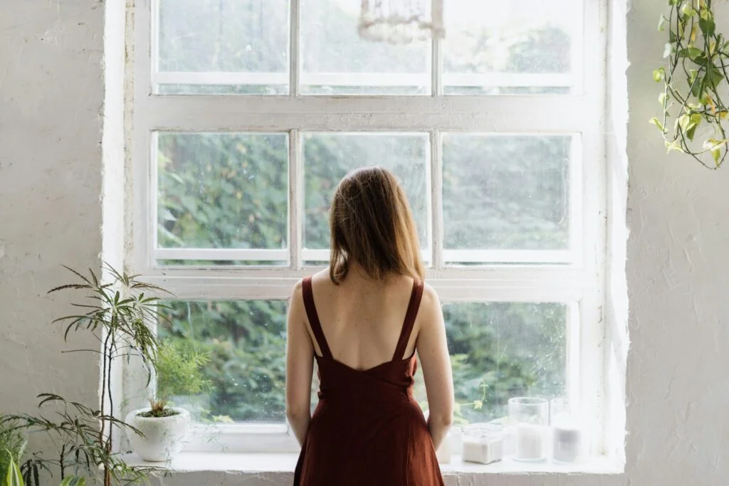 A woman in a red dress standing at a large window looking out at green trees, lost in thought