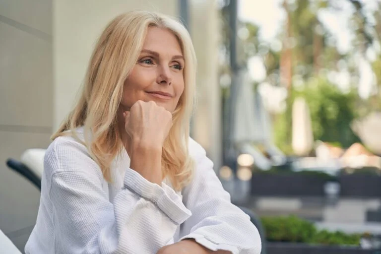 A smiling woman in her fifties in a white top resting her chin on her hand outdoors