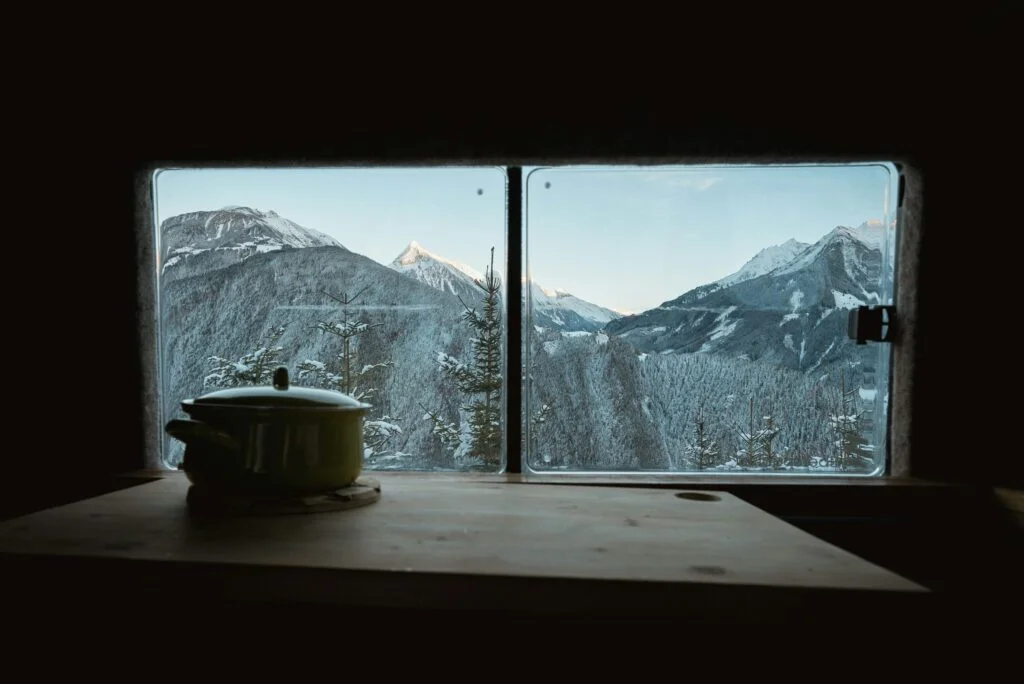 A dark room looking out through a window at snowy mountains on a winter morning