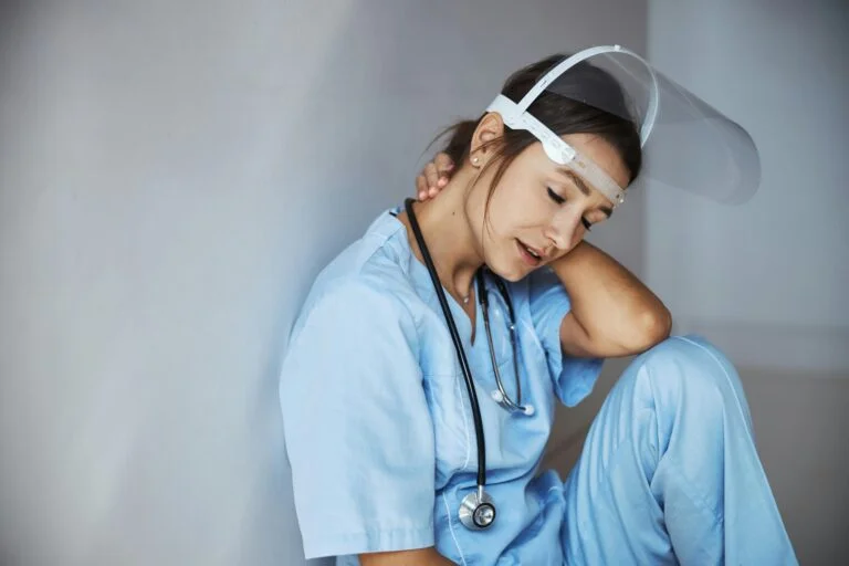 A tired nurse in scrubs and a face shield resting against a wall after a shift