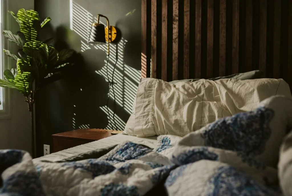 A calm bedroom with a made bed, a plant and soft morning light through the blinds