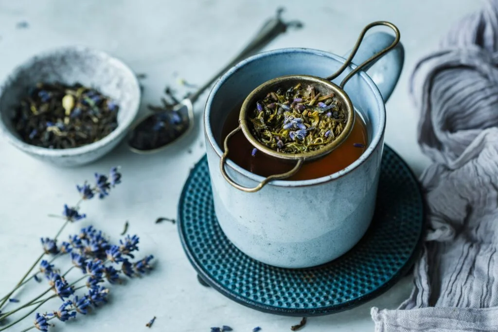 A mug of herbal tea with a strainer of dried lavender and herbs on a blue coaster