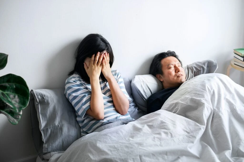 A woman awake in bed covering her face while her partner snores beside her