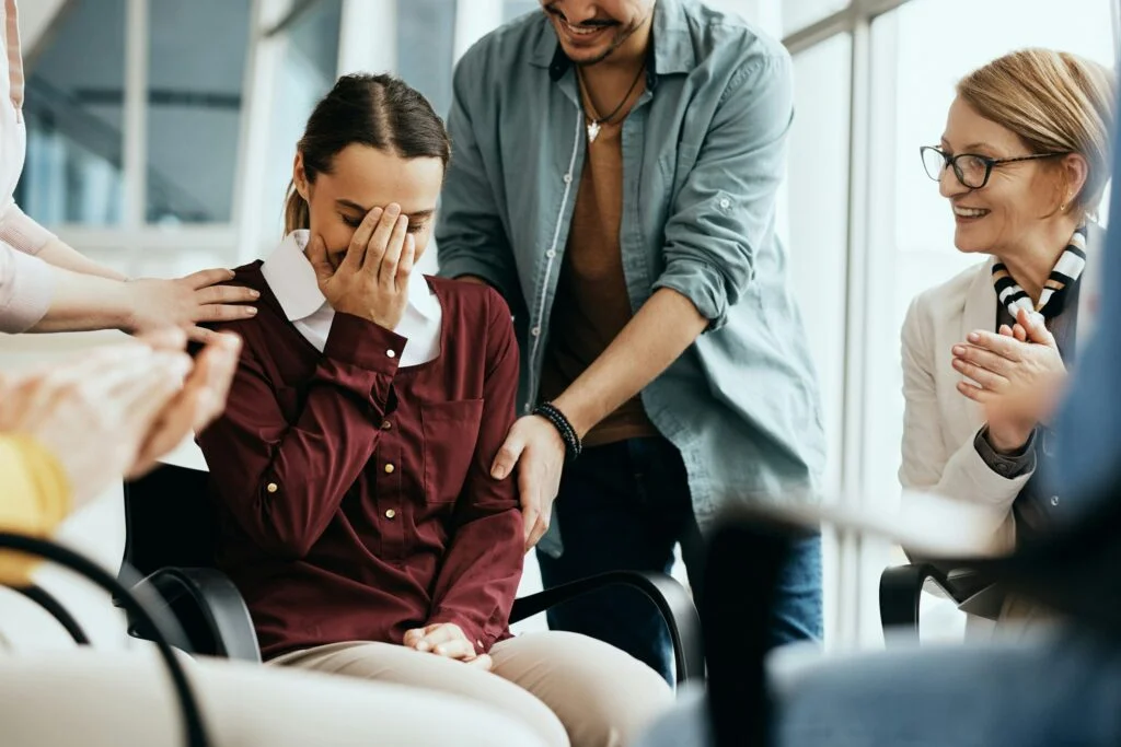 A woman covering her face with her hand while people around her smile and clap