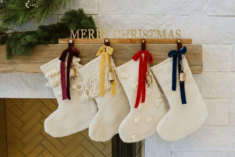 Four cream knitted Christmas stockings with coloured ribbons hanging from a mantelpiece