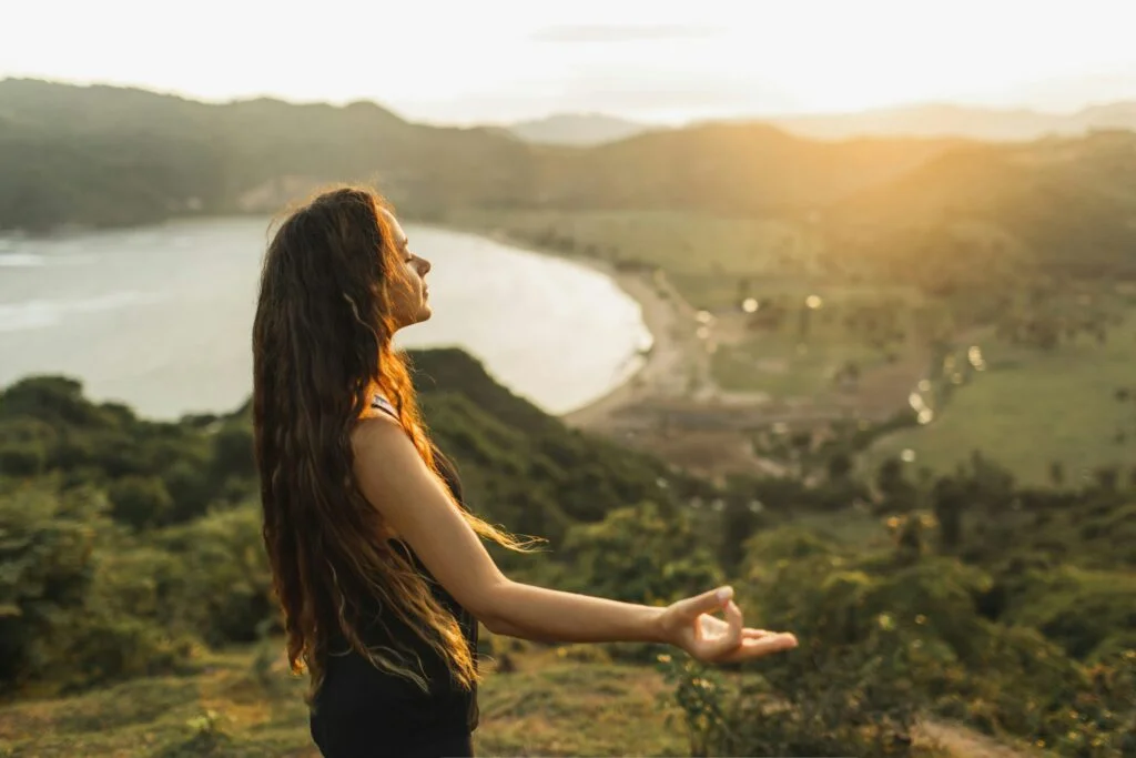 A woman with long hair breathing calmly with her eyes closed on a hillside above the sea at sunset