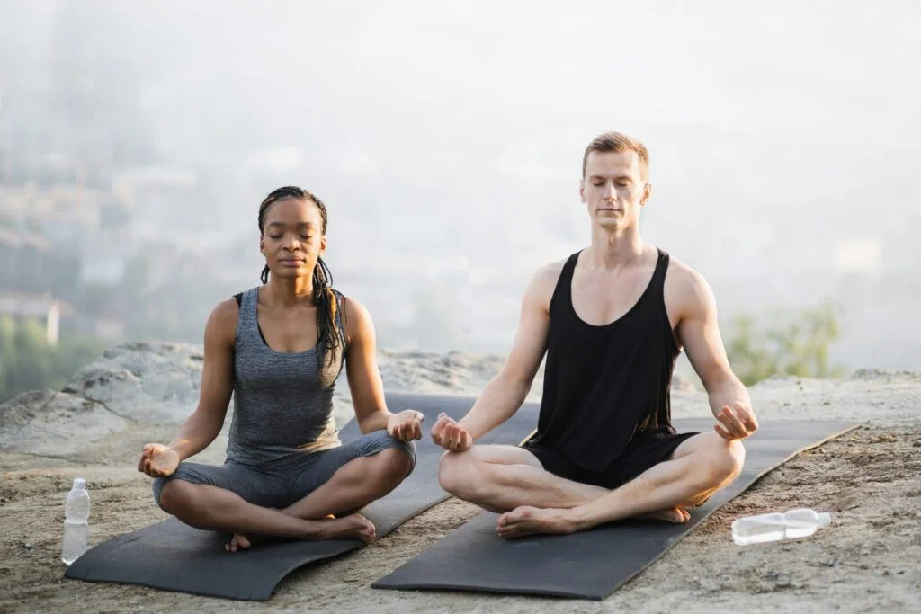 Two people meditating cross-legged on mats outdoors on a misty morning
