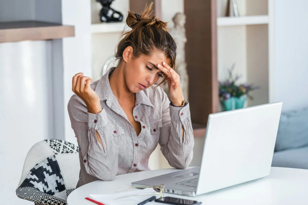 A stressed woman at a desk holding her forehead in front of a laptop
