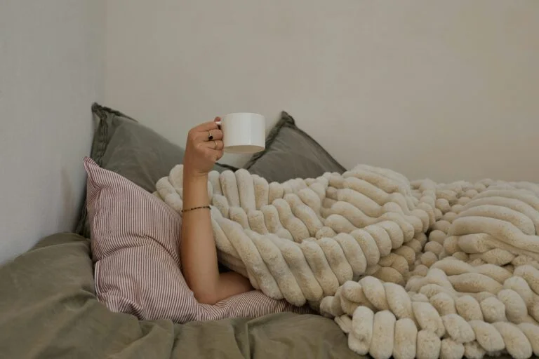 A person under a chunky knitted cream blanket in bed holding up a white mug
