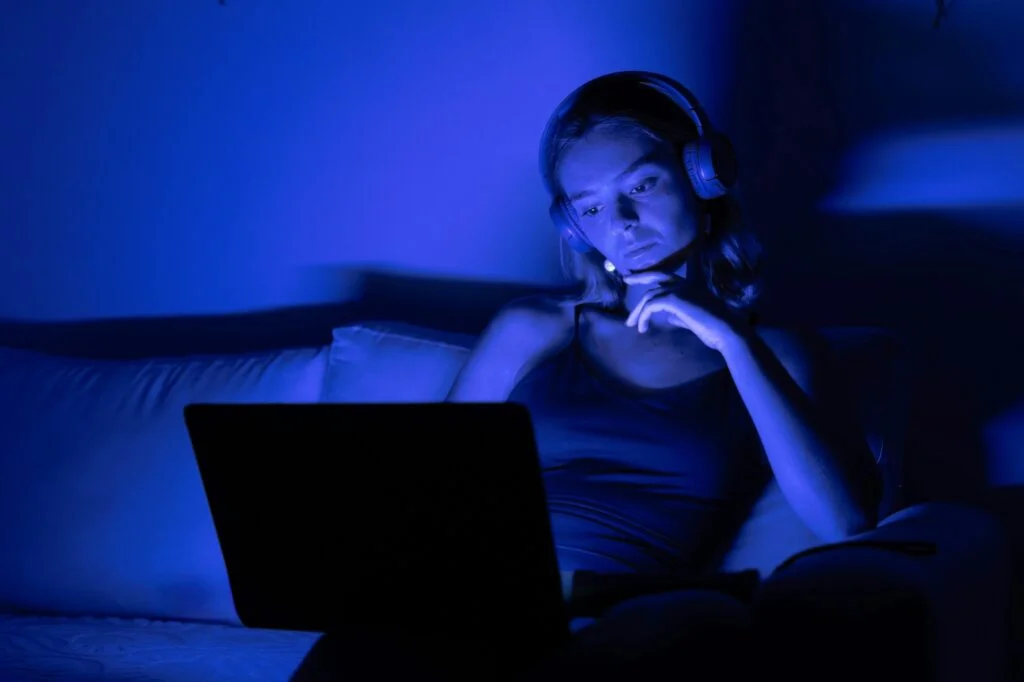 A woman with headphones looking at a laptop in a dark room lit by blue screen light