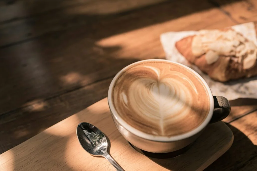 A latte with heart-shaped foam art next to a croissant on a sunlit wooden table