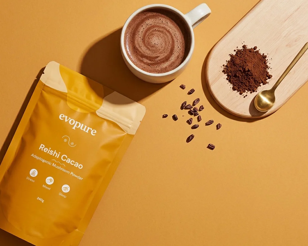 A pouch of Evopure Reishi Cacao beside a mug of hot chocolate, cacao powder on a wooden board and scattered cacao beans