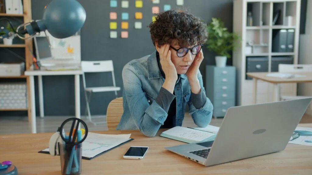 A woman at her desk holding her head in her hands in front of a laptop