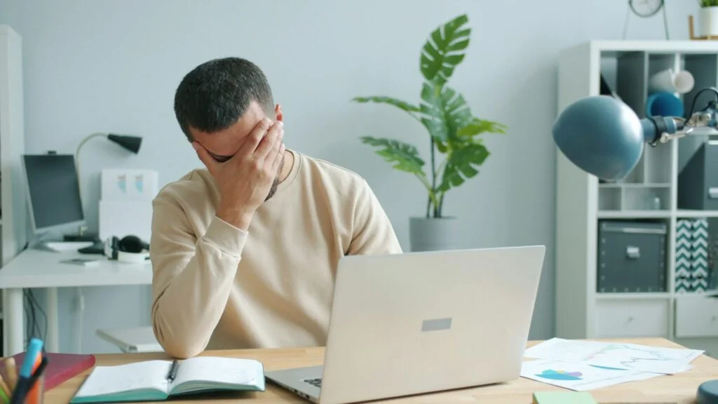 A man at his desk rubbing his face in front of a laptop, looking stressed