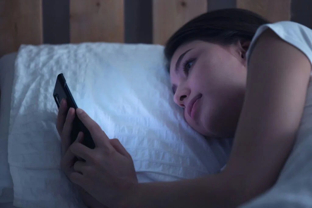 A young woman lying in bed at night scrolling on her phone