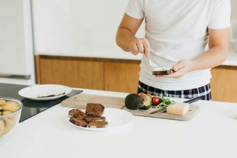 A man in pyjamas spreading cheese on toast in the kitchen with avocado and tomatoes nearby