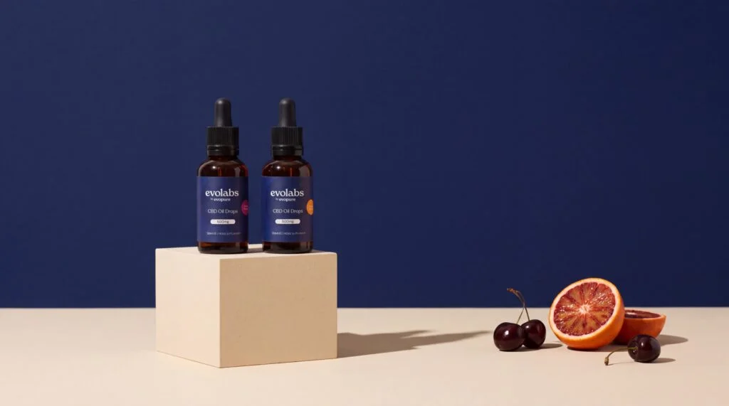 Two Evolabs by Evopure CBD oil bottles on a plinth with cherries and blood orange on a navy background