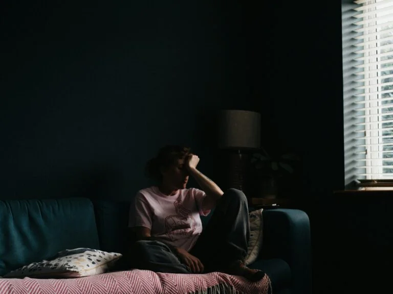 A person sitting on a sofa in a dim room, resting their head on their hand