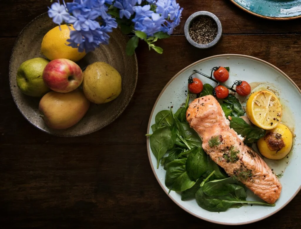 A plate of salmon with spinach and tomatoes next to a bowl of fresh fruit