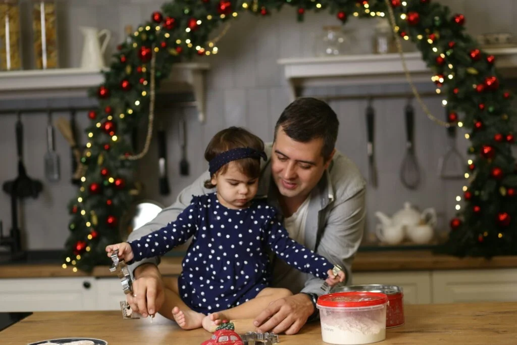 A dad and his young daughter cutting out Christmas biscuits together at a kitchen table under a festive garland