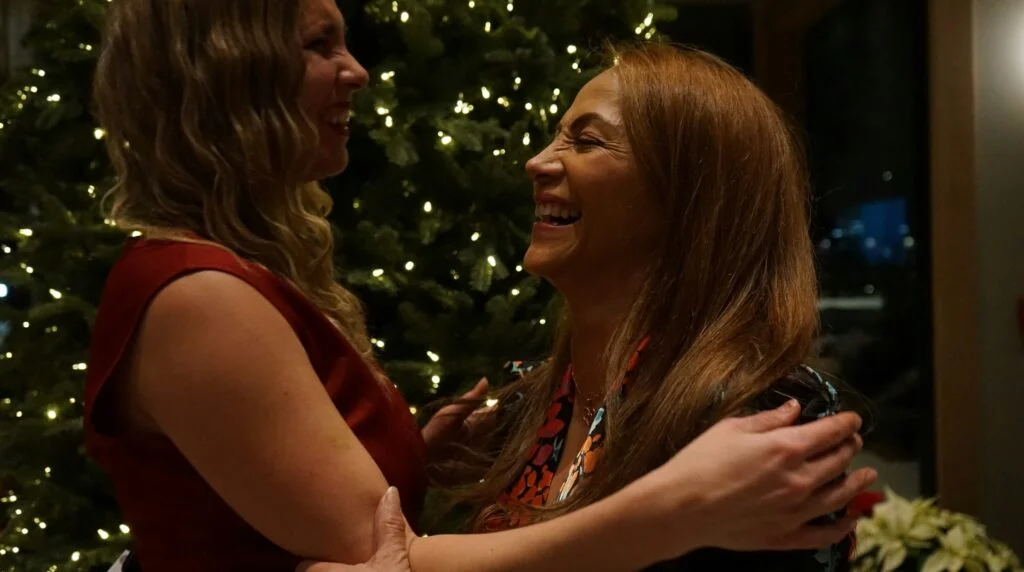 A smiling mother and her adult daughter hugging in front of a lit Christmas tree