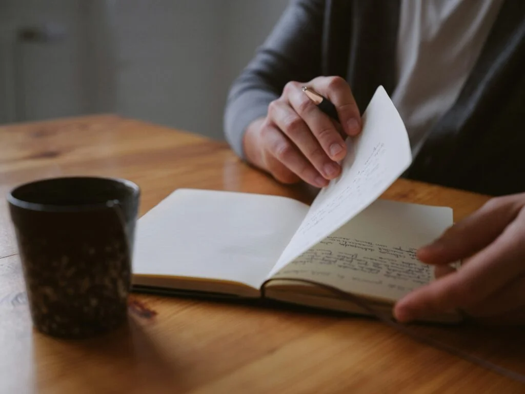 Hands turning the pages of a handwritten journal beside a mug on a wooden table