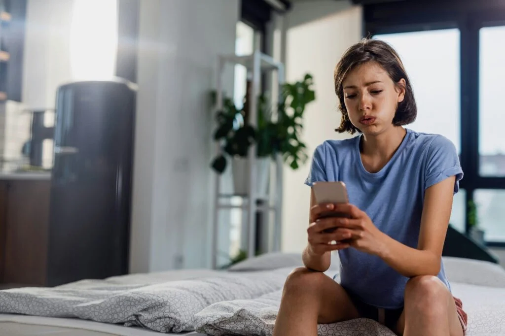 A worried young woman sitting on her bed looking at her phone