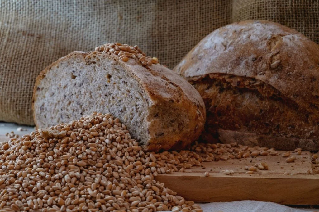 A halved loaf of wholegrain bread beside a pile of wheat grains