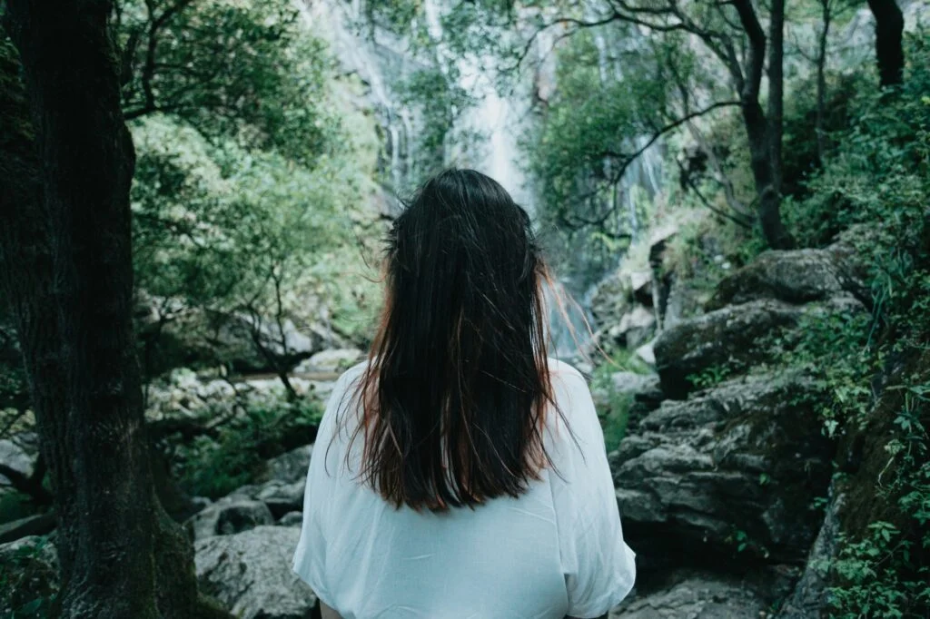 A woman with long dark hair standing quietly in a green forest by a waterfall