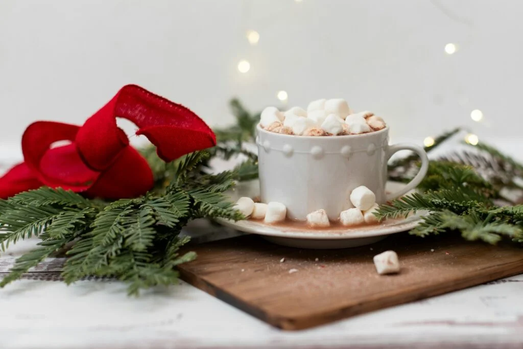 A cup of hot chocolate topped with mini marshmallows on a wooden board with pine sprigs and fairy lights