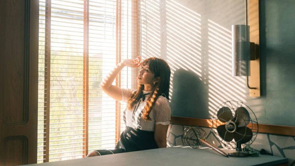 Woman by a sunny window holding her forehead beside an electric fan