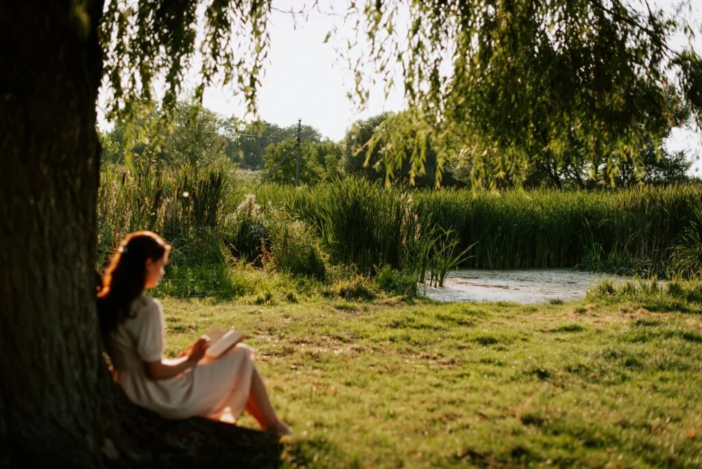 A woman reading a book under a willow tree beside a pond on a calm summer evening